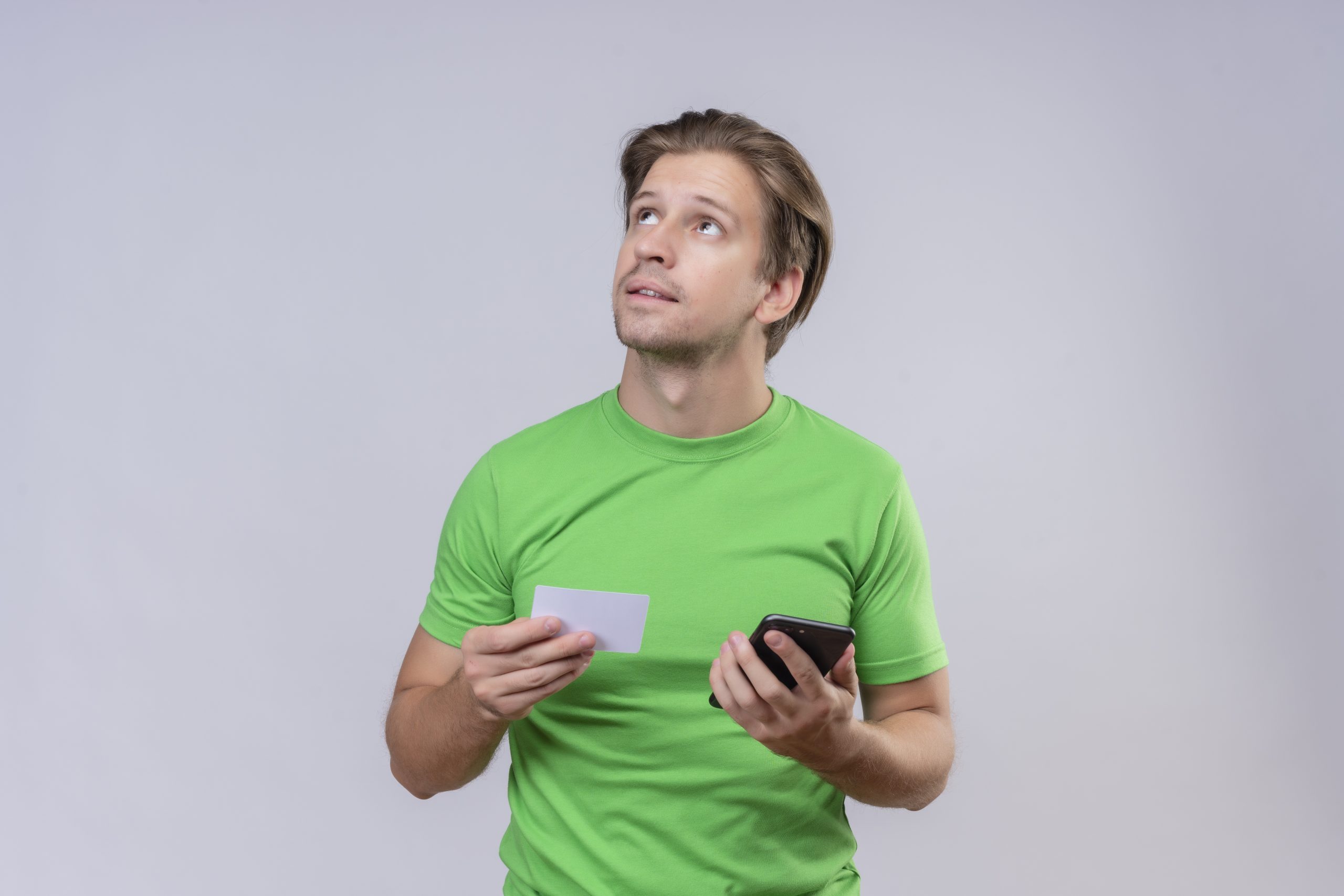 young handsome man wearing green t-shirt holding smartphone and credit card looking up with pensive expression on face thinking trying to make choice standing over white background